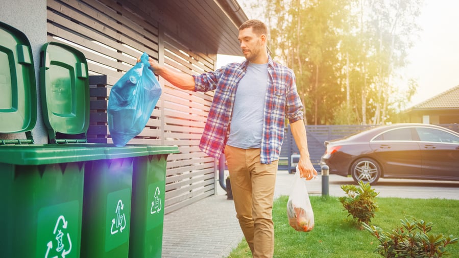 A Man Is Throwing Away Two Plastic Bags Of Trash Next To His House