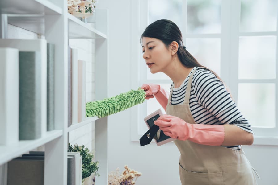 Housekeeper In Apron Dusting The Bookshelf By Feather Duster