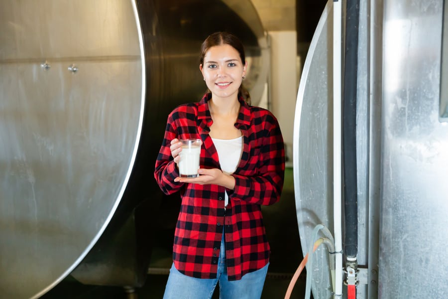 Smiling Young Workwoman Standing Near Large Tanks In Storage Room Of Dairy Farm, Holding Glass With Fresh Milk