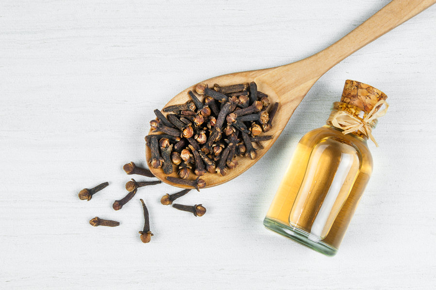 Close Up Glass Bottle Of Clove Oil And Cloves In Wooden Spoon On White Rustic Table.