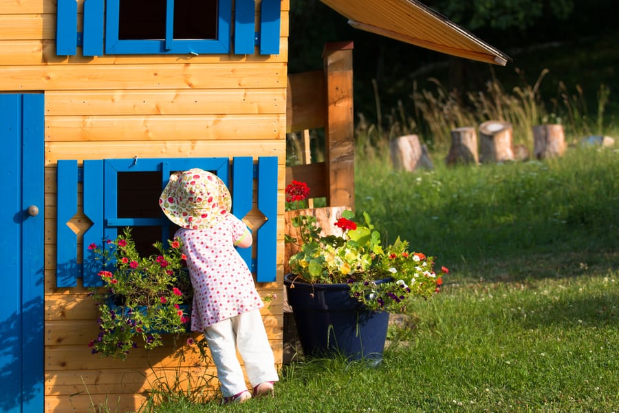 Little Cute Curios Girl Looking Into The Blue Window Of Wooden Playhouse In The Countryside Garden With Blooming Flowers