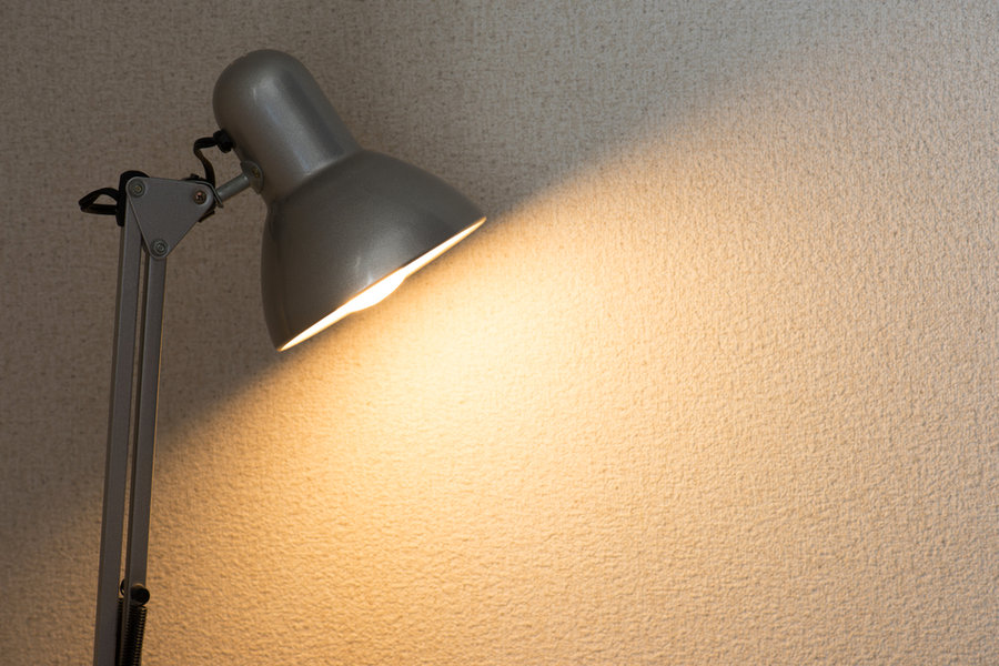 Silver Desk Lamp Or Arm Lamp In Front Of A White Wall With Incandescent Type Light Bulb Illuminating The Wall.