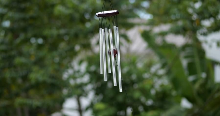 Wind Chimes Hangs On The Porch Of The House Against The Backdrop Of Green Trees