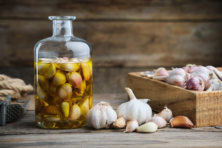 Garlic Aromatic Flavored Oil Or Infusion Bottle, Wooden Crate Of Garlic Cloves And Garlic Press On Wooden Kitchen Table.