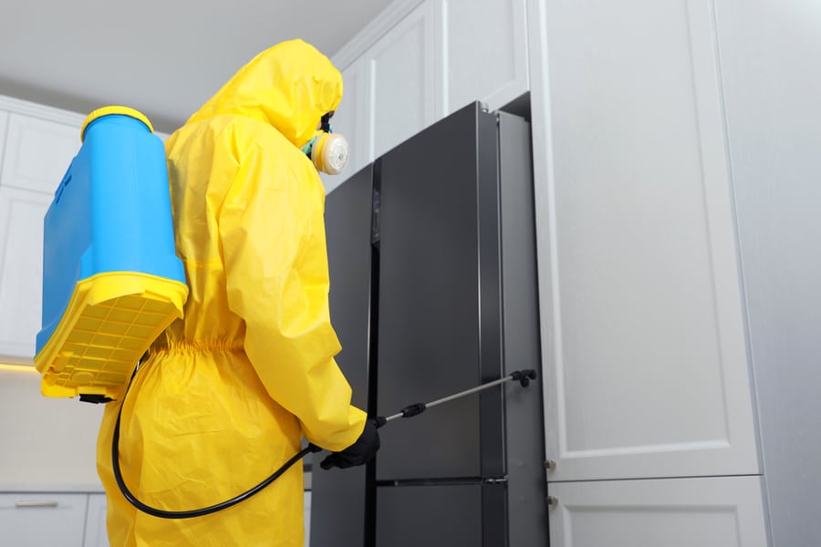 Pest Control Worker In Protective Suit Spraying Insecticide Near Refrigerator Indoors